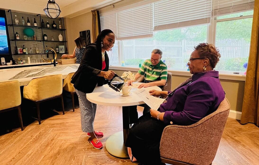 Caregiver serves coffee to residents sitting at a round table in a community lounge.