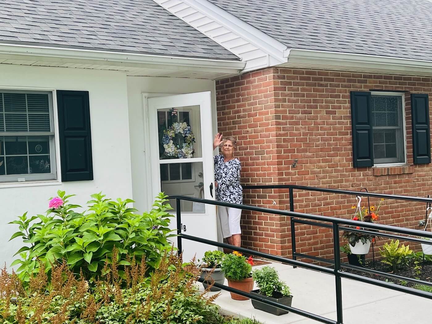 An elderly woman waves from the entrance of a brick unit with black shutters and a ramp.