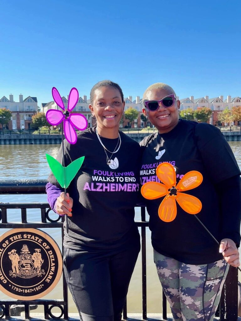 Two people holding flowers at a walk event near a river with residential units behind them.