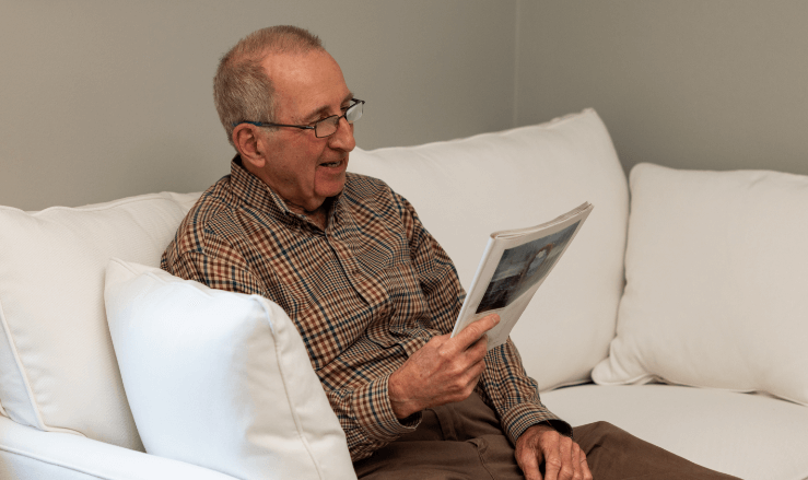 Smiling elderly man relaxing on a couch reading a magazine in a cozy unit.