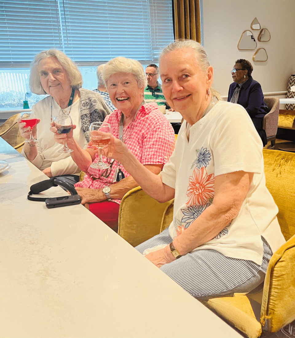 Three women smiling and toasting with drinks in a community gathering area.