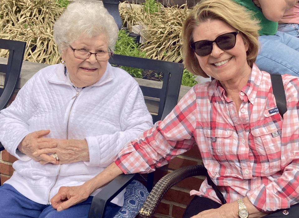Two women, one elderly, sitting outdoors holding hands and smiling at a senior living community.