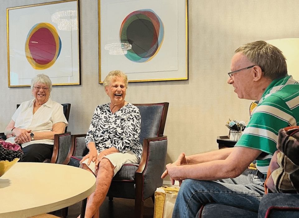 Three seniors enjoy a lively conversation in a warmly decorated community lounge.